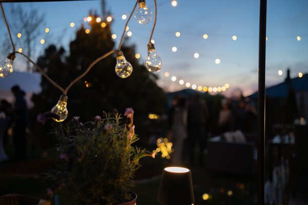 Lichterketten und Glühbirnen in der Abenddämmerung, im Hintergrund Gäste einer Gartenfeier