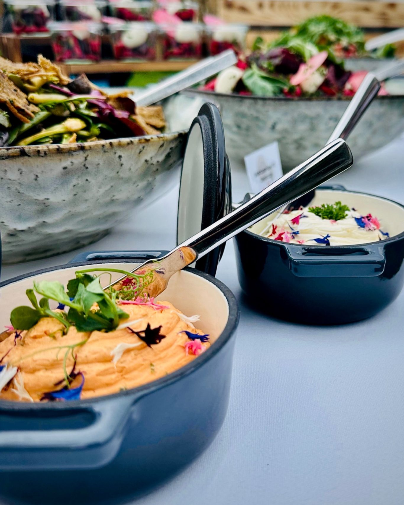 Zwei Dips in gusseisernen Töpfchen mit essbaren Blüten, dahinter Salatschalen auf der Buffettafel